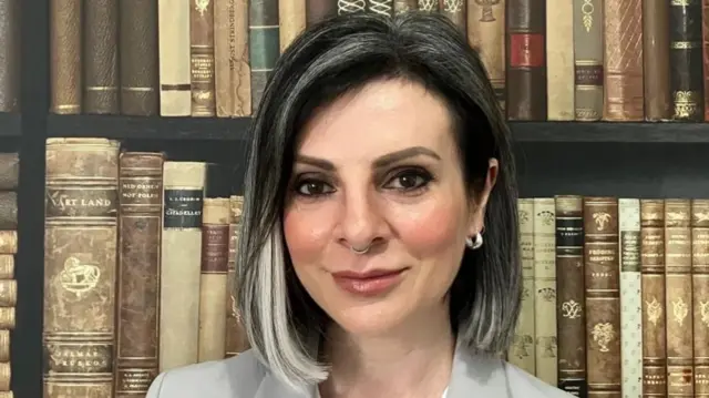 Lucy Cohen looks into the camera while stood in front of a bookshelf full of old books. She has black hair with white streaks and a nose ring