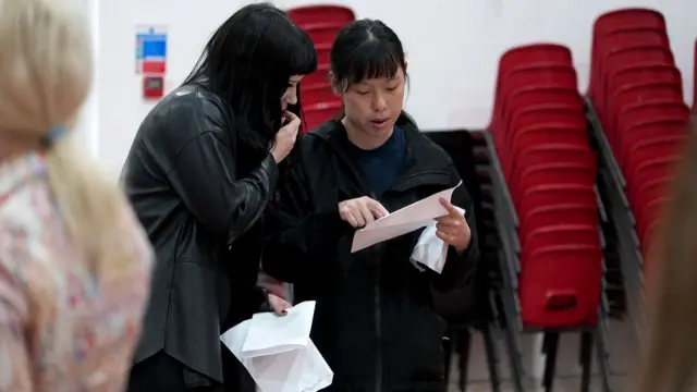 Two female students with dark hair and wearing dark clothing are looking over their results sheets in a school hall