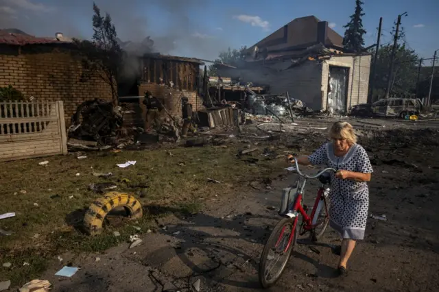 Several buildings damaged after Russian shelling in eastern Ukrainian town of Pokrovski, with firefighters attending a house with smoke as a woman walks past with a bicycle