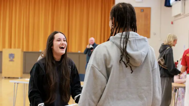 A girl in the background with long dark hair smiles at a friend, who has their back to camera wearing a grey hoodie