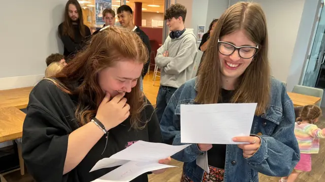 Two girls look at their results sheets. The one on the left has red hair and is wearing a dark hoodie, on the right she is wearing a denim jacket and has glasses