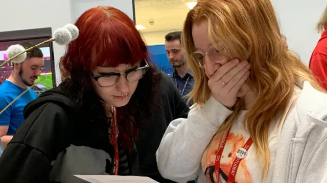 Two students look over one of their results papers. The girl on the right, wearing a light grey hoodie, has her hand clasped over her mouth