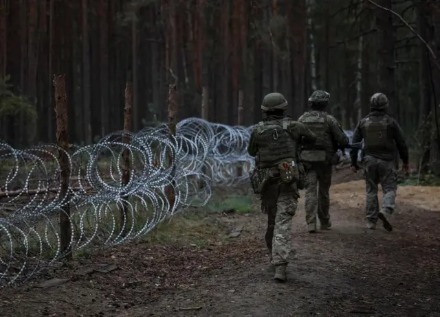 Three Ukrainian soldiers, in full gear and with their backs facing the camera, on patrol near the border with Belarus in September 2023, the border marked with barbed wire