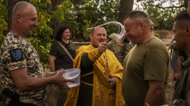 An army chaplain flick water from the end of a brush at a soldier, who braces for the water to hit his face. A soldier next to the chaplain holds a clear plastic tub with water in it