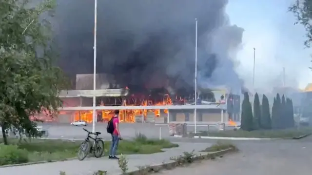 A building is on fire with lots of black smoke rising from it. A man wearing a red t shirt and black backpack looks on at the fire while stood next to a bike