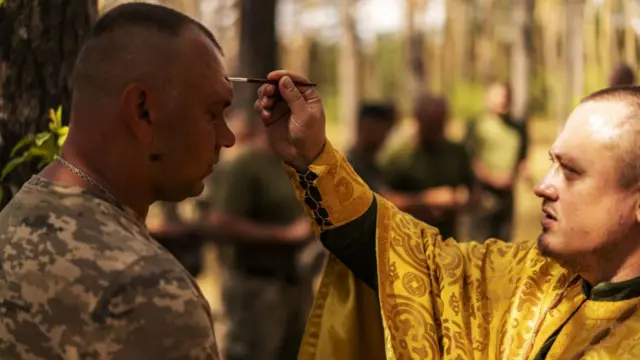 An army chaplain holding a thin paintbrush wipes it across a soldier's head