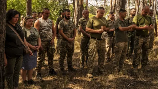 Ukrainian soldiers, dressed in dark green and camouflage clothing, stand in a forest with each of them holding their own hands together in front of them