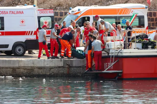 Around a dozen emergency services worker on a quay, as a body bag is brought ashore from a boat