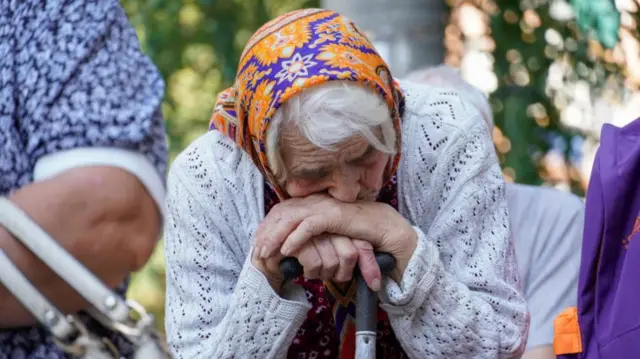 An elderly woman with a scarf tied around her head leans her head on her cane as she is being evacuated from the Kursk regional border with Ukraine