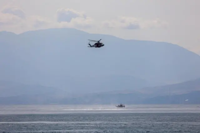 A helicopter flies overhead as a rescue boat moves on the surface of the sea, a large mountain is visible in the backdrop