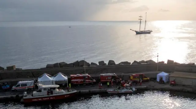 A aerial view shows emergency and rescue service vehicles and personnel at a port, a different yacht is visiible on the open sea behind them