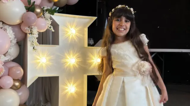 Alice da Silva Aguiar smiling, she is wearing a white dress and standing next to a cross with lights on it