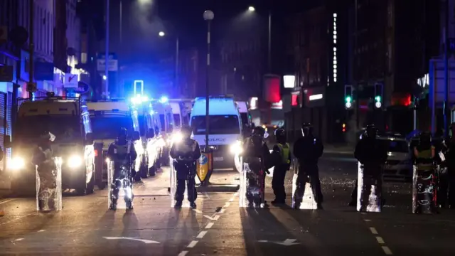 Seven Merseyside Police officers in riot gear, waring helmets and holding shields in front of their legs, stand in a line in Liverpool. At least six police cans are parked in front of them.