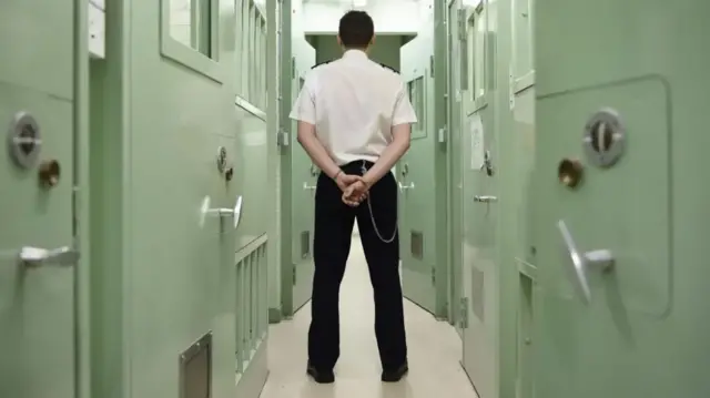 A prison guard stands in the middle of a hallway of prison cells with his hands folded behind his back