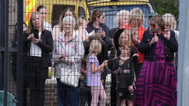 A group of women and children wait for King Charles behind a wire fence