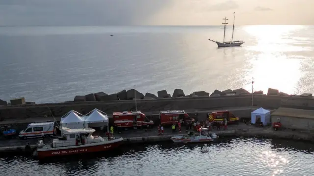 Sheltered behind a constructed rock wall on the water, tents are erected and ambulances and boats gather