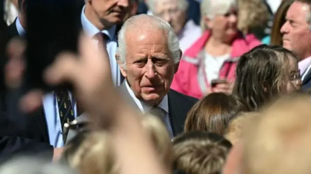 King Charles in a crowd of people. Some in the foreground are taking pictures of him on their phones.