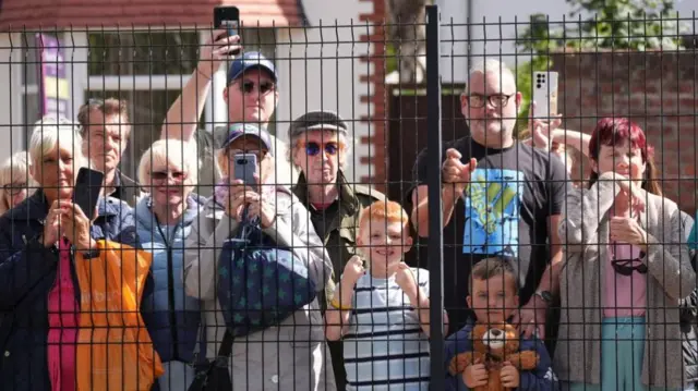 A group of pople and children wait for King Charles behind a wire fence