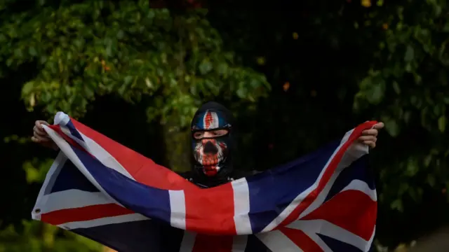 A man with his face covered holds up a union jack flag