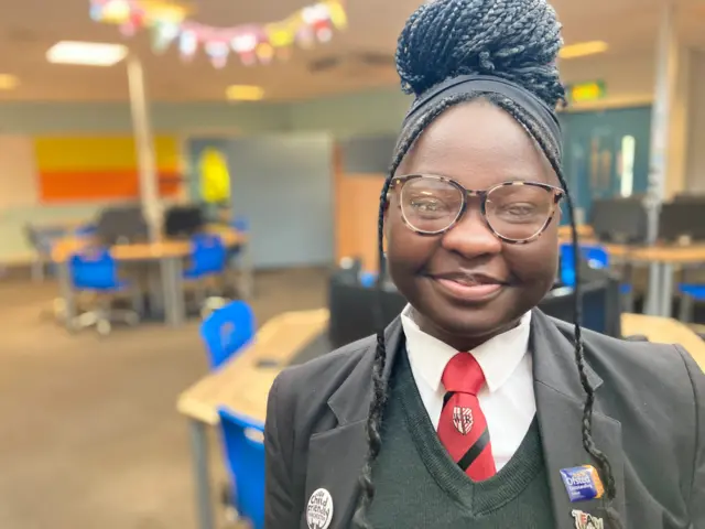 Cheryl is stood in a classroom and smiling at the camera. She wears a white shirt, red school tie, black sweater and a blazer with colourful badges on it. She has black hair tied up in a bun