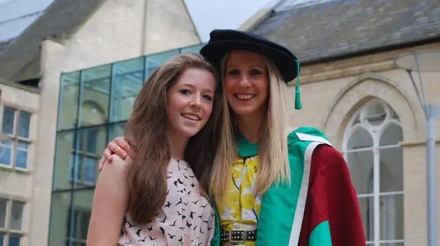 Anna smiles at the camera, wearing a green and red graduation gown and a yellow dress. She has her arm round her daughter Star, who is wearing a patterned light pink dress.