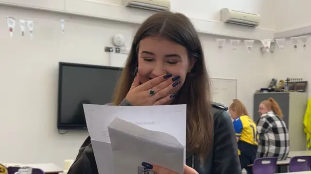 A girl smiling as she reads from a piece of paper, her hand is covering her mouth