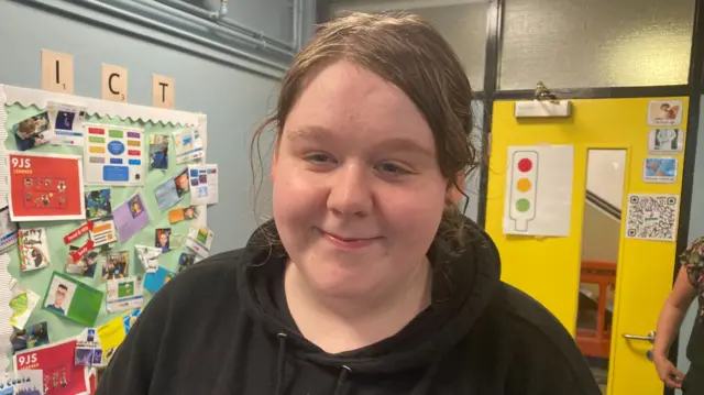 Cobhlith has long hair tied back in a ponytail and is wearing a black hoody. She's stood in front of a colourful classroom display, and a bright yellow classroom door