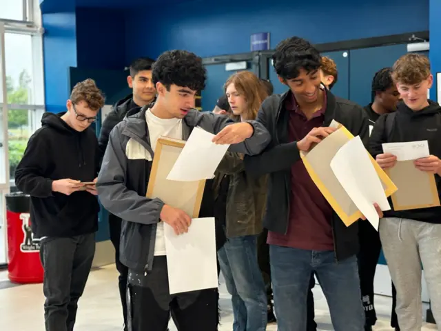 Two male students wearing jackets in the foreground open their results in the lobby area of their school. One is smiling looking into the envelope and the other is looking at his sheet open-mouthed