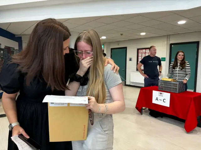 A girl with long blonde hair and glasses holds her open results envelope in front of her, with her hand over her mouth and tears in her eyes. A lady with dark hair is embracing her and smiling