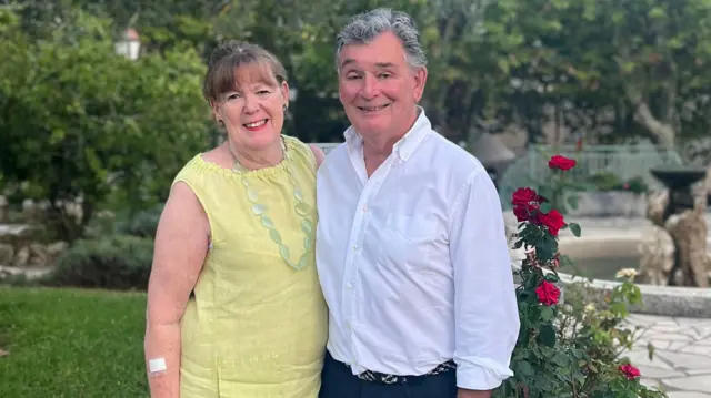 Judy Bloomer (left) and Jonathan Bloomer (right) pictured side by side with arms around each other in formal attire surrounded by cobbled paving and flowers in an outside garden