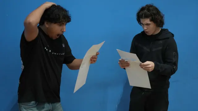 Two boys stand with shocked expressions as they read their GCSE results