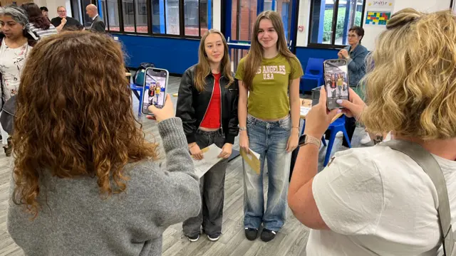 Two girls pose for a photo being taken by two women on their phones