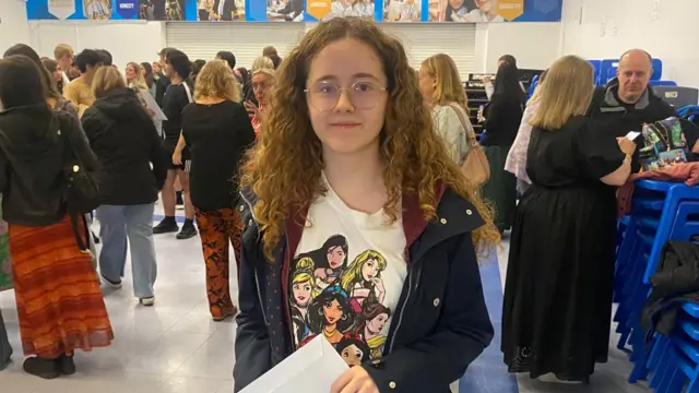 A girl holding an envelope containing her GCSE results. She is wearing glasses and a white T-shirt, there are lots of people in the background