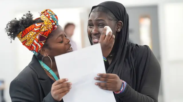 Girl wipes her tears smiling with a tissue while holding her GCSE results, a woman stands next to her