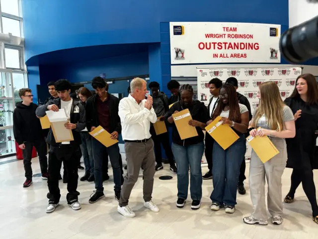 A large group of students open their results envelopes with a man in a white shirt clapping in front of them