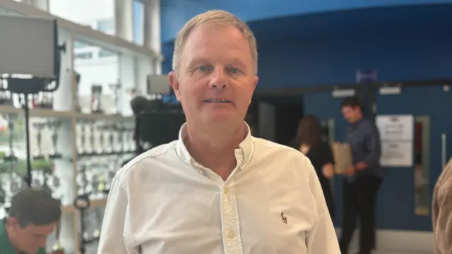 Martin Haworth, wearing a white shirt, looks into the camera from the lobby area of his school