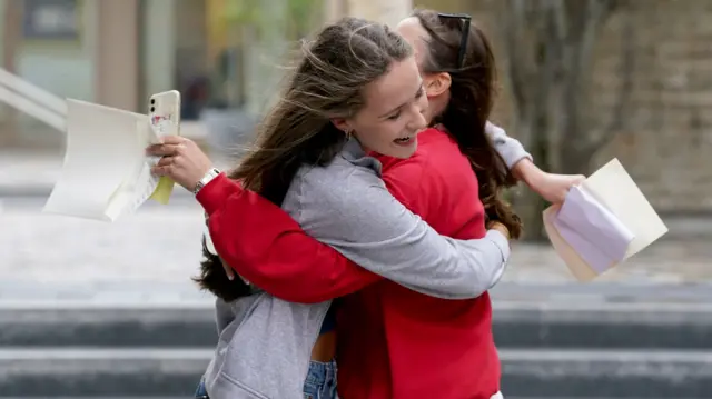 Two girls hug each other while holding pieces of paper with their GCSE results on them