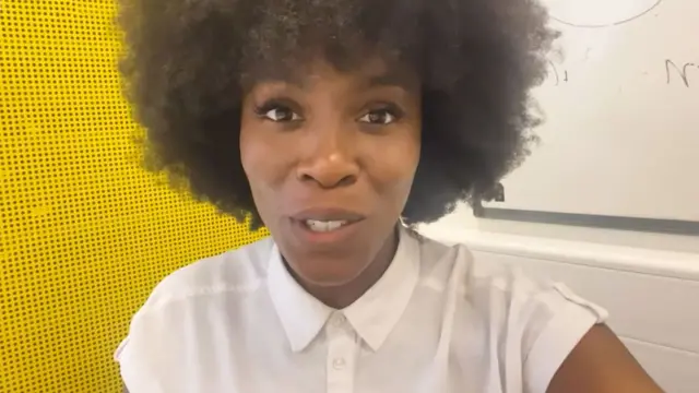 Secondary school teacher Youlande is speaking into the camera. She has afro hair and is wearing a white short-sleeved shirt. She is standing in front of a whiteboard in a classroom and a bright yellow wall display.