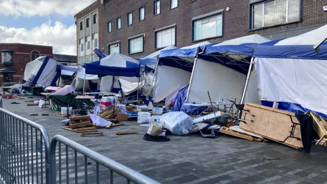 Blue and white market stalls behind a grey fence. Gazebos have been blown backwards and the contents of them are scattered across the street infront