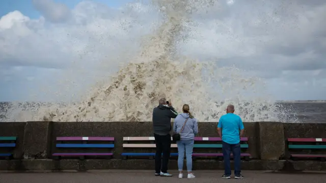 Three people stand behind sea wall, with one taking a picture with a camera, while wave crashes into the sea wall and up into the air