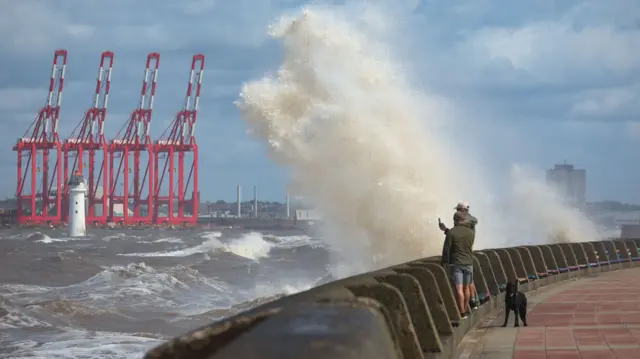 Two people stand next to a dog as they lean on sea wall as water sprays into the sky