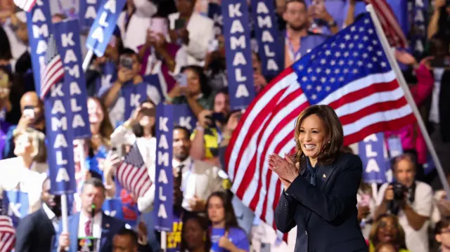 Kamala Harris claps her hands together in front of a large crowd with an American flag in the background