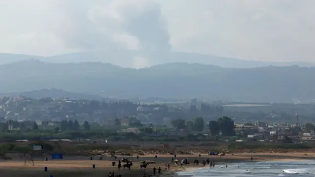 A view shows smoke on the Lebanese side of the border with Israel, as people walk along a beach in Tyre, Lebanon (25 August 2024)