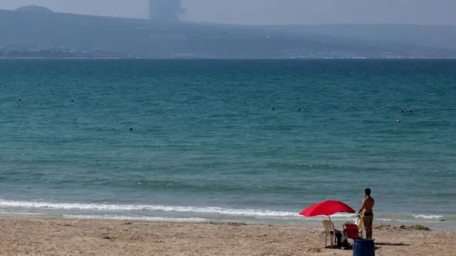 A man stands at a beach in Tyre, Lebanon, looks at smoke rising in the distance