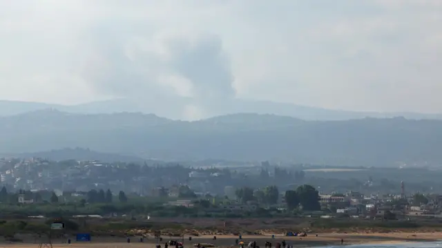 A view shows smoke on the Lebanese side of the border with Israel, as people walk along a beach in Tyre