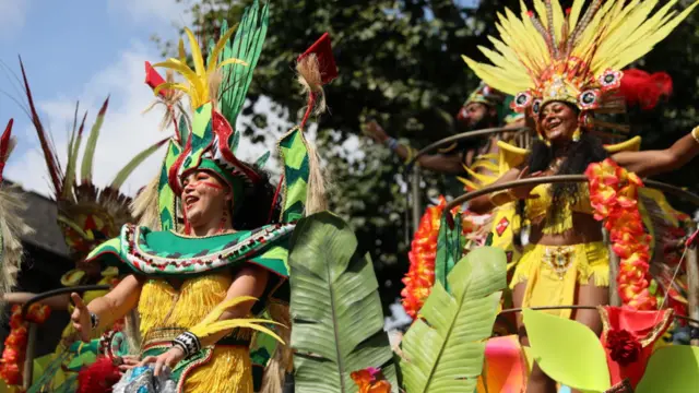 Two women in brightly coloured outfits in the parade with feathers on their outfits, leaves and moving floats at different heights