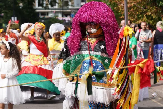 A man in a sparkling costume. He has a purple wig and a custom-made top with the Brazilian flag on the front