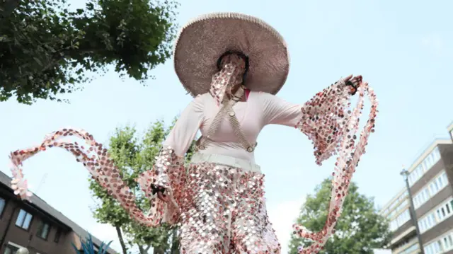 A woman in pink outfit with sequin trousers, sleeves, hat and a piece of sequin fabric covering her face