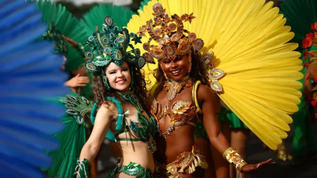 Two women at Notting Hill Carnival in brightly coloured yellow and green outfits with large fan-like structures on their back in bright colours and jewelled headpieces to match their outfits