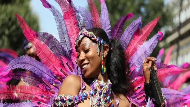 Woman in purple and pink outfit at Notting Hill Carnival with large feathers in a semi-circle behind her